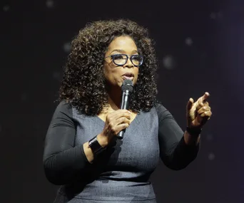 Oprah Winfrey speaks on stage, holding a microphone and gesturing with her hand.