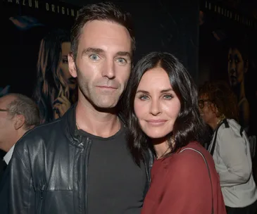 Couple posing together at an event, with posters visible in the background.