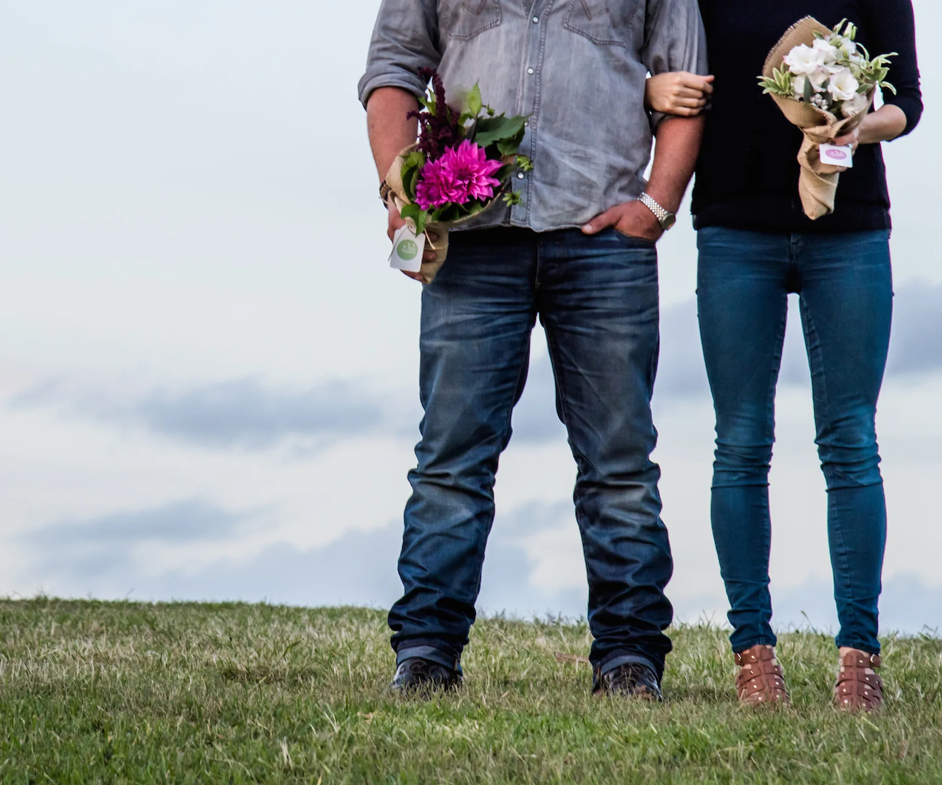 A couple stands arm-in-arm holding bouquets, against a cloudy sky backdrop.