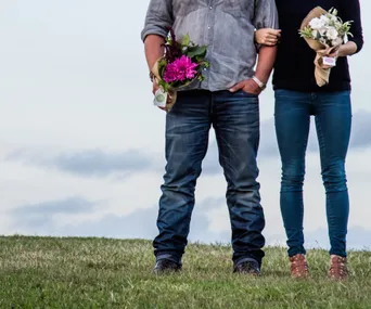 A couple stands arm-in-arm holding bouquets, against a cloudy sky backdrop.