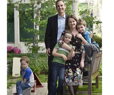 Family photo with four people posing happily and a child sitting apart, outdoors.