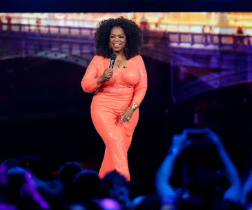 A person in a coral dress speaks on stage holding a microphone, with a crowd and vibrant background behind.