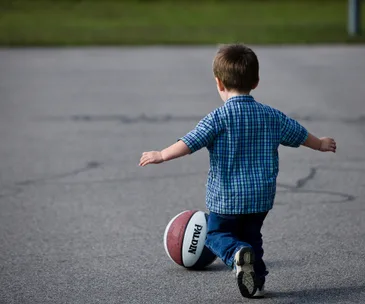 Child playing basketball outside on pavement, wearing a blue checked shirt and jeans.