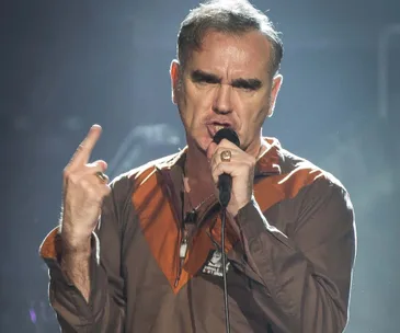 A man singing passionately into a microphone on stage, wearing a brown and orange jacket.