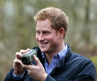 A smiling man holds a camera against a blurred outdoor background.