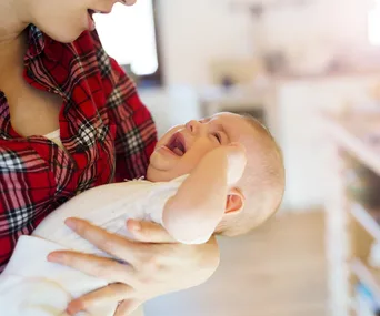 A person in a red plaid shirt holds a smiling baby dressed in white, in a softly lit room.
