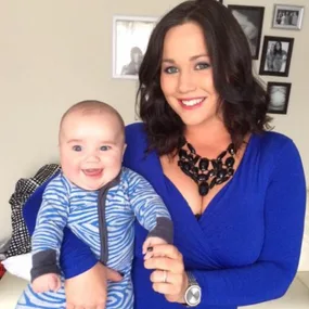 A woman in a blue dress smiles while holding a baby in a two-tone onesie, standing in a room with framed photos.