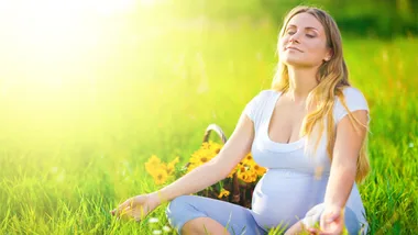 Pregnant woman sitting in a sunny meadow, eyes closed and smiling, surrounded by flowers.