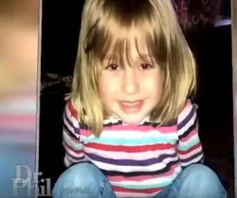 Young child with blonde hair, smiling, wearing a striped shirt and sitting cross-legged on a Dr. Phil show screenshot.