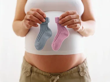 Pregnant woman holds one blue and one pink baby sock, symbolizing gender selection or baby sex prediction.