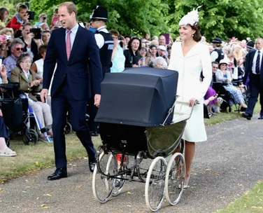 A couple walking with a vintage baby pram in a crowd; both dressed formally, surrounded by onlookers.