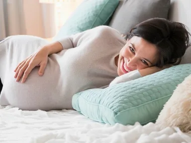 Pregnant woman smiling, lying on bed with teal pillows, hand on belly, wearing a light sweater.
