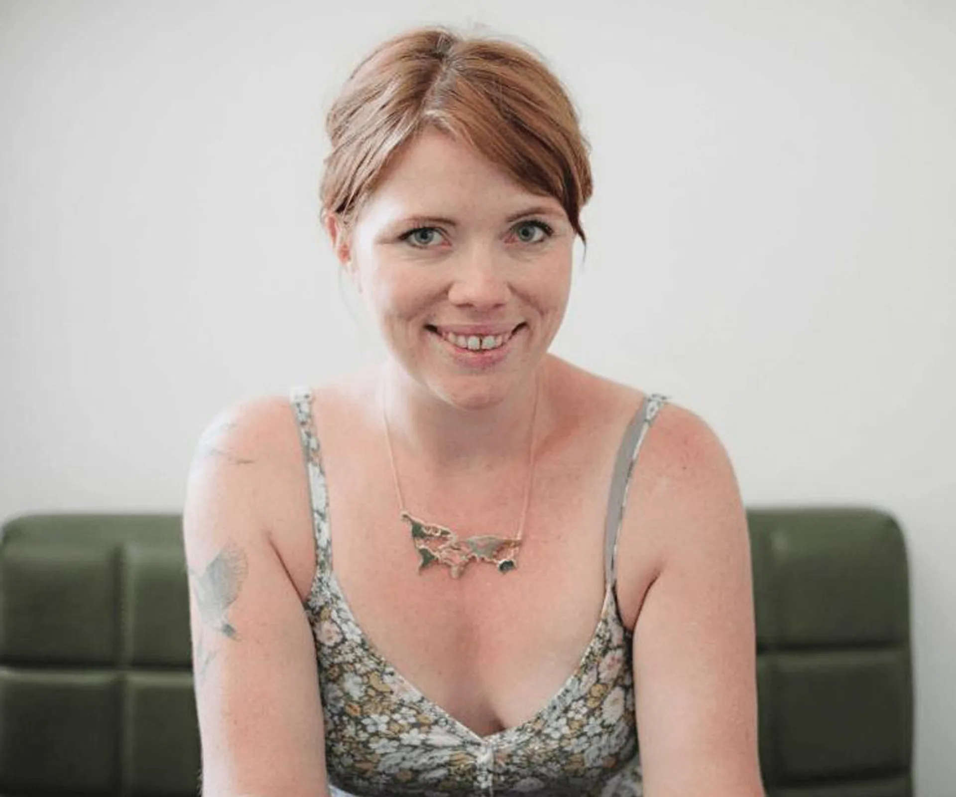 A woman with red hair smiles, wearing a floral top and necklace, seated indoors.