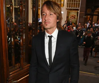 Man in a black suit and tie stands in front of a wooden door amidst a crowd on a city street.