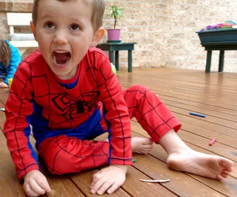 A child in a Spider-Man costume sitting on a wooden deck, appearing excited or playful, with crayons scattered around.