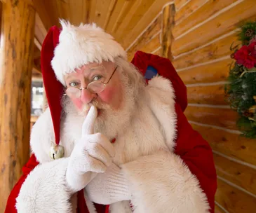 Santa Claus in red suit and hat, with finger over lips, shushing. Background shows wood paneling and greenery.