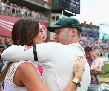 A cricketer in a cap hugs a woman, with spectators in the background at a sports stadium.