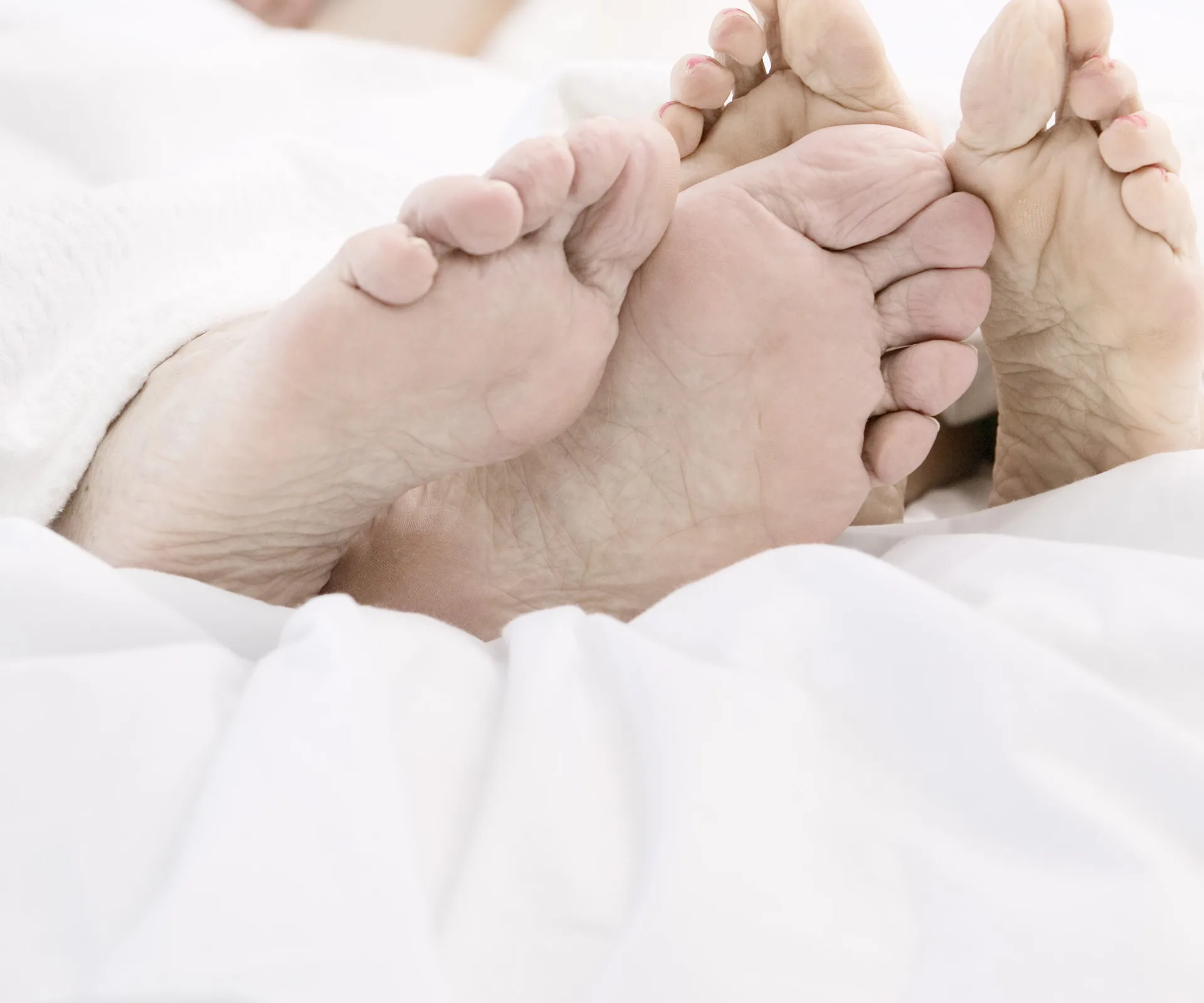 Two pairs of feet intertwined under white bedsheets.