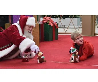 Santa Claus plays on the floor with a young boy holding a toy, surrounded by holiday decorations and gifts.