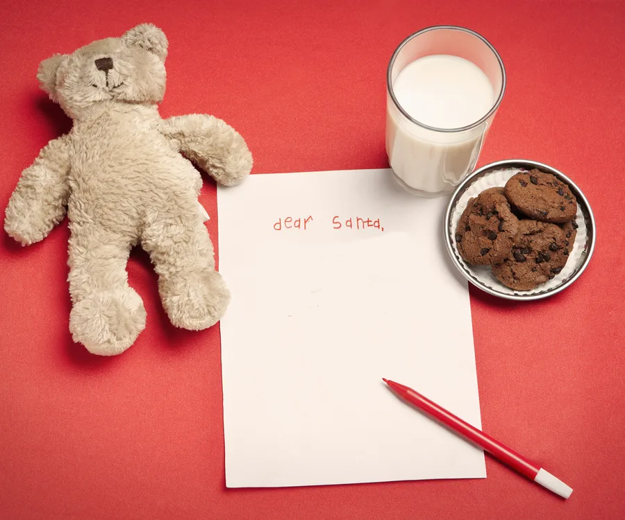 Teddy bear, letter to Santa, red pen, glass of milk, and plate of cookies on a red background.