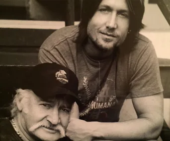 Black and white photo of two men sitting, one in a cap and the other with long hair, casually posing for the camera.