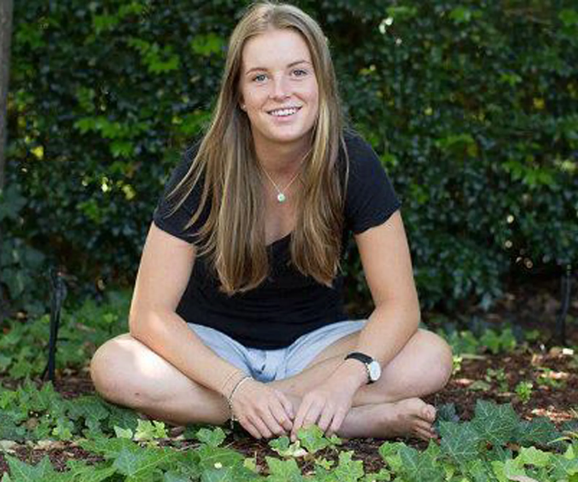 A young woman sits cross-legged on the ground outdoors, surrounded by greenery, smiling at the camera.