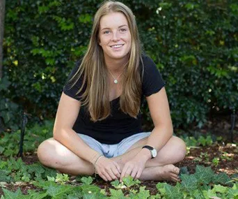 A young woman sits cross-legged on the ground outdoors, surrounded by greenery, smiling at the camera.