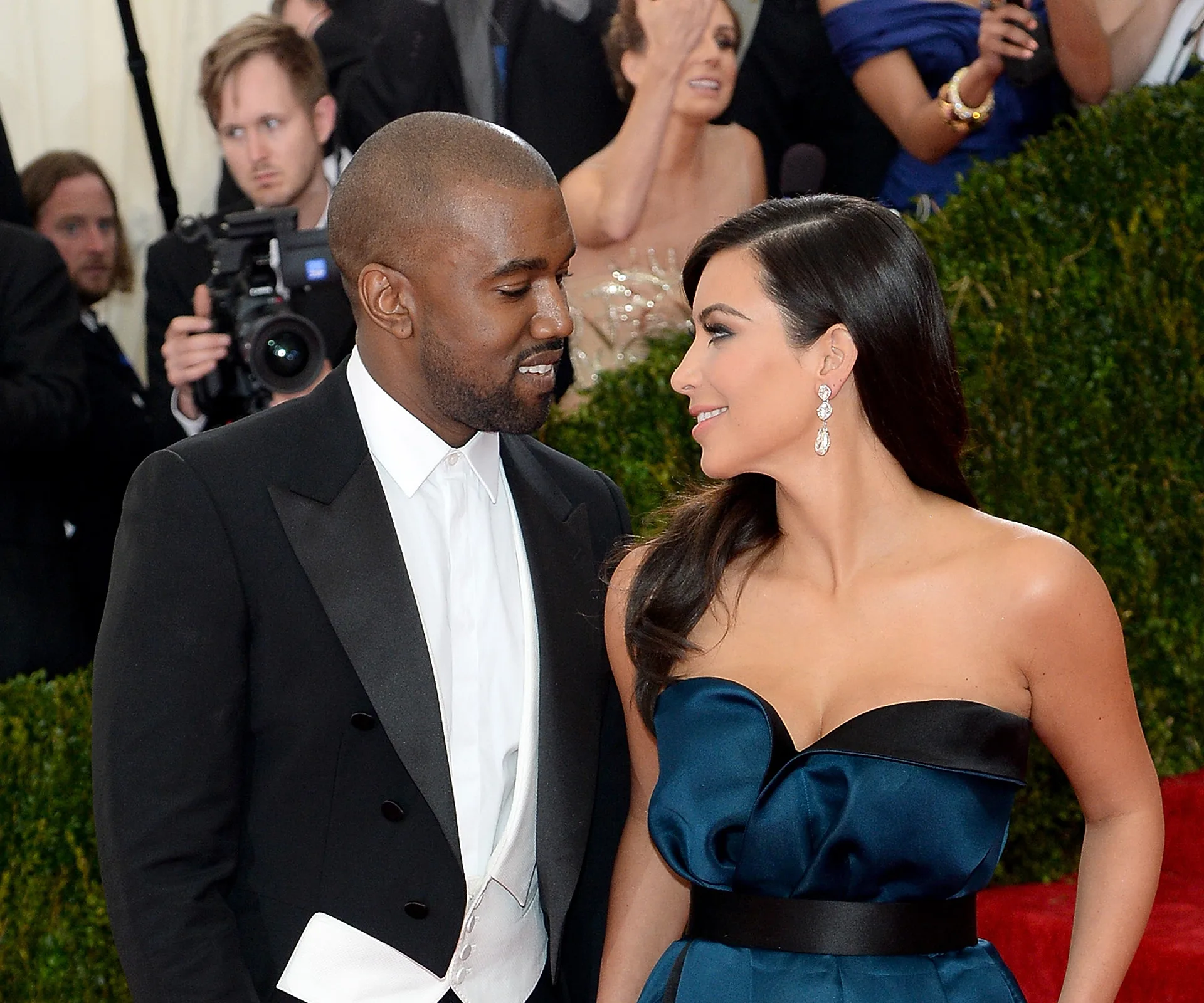 Kim Kardashian and Kanye West smiling at each other on a red carpet event, surrounded by photographers.