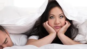 A couple lying in bed, woman looking frustrated under a blanket with her hands on her cheeks.