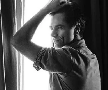 Man posing by a bright window, gazing outside, with hand in hair, exuding a relaxed and thoughtful vibe.