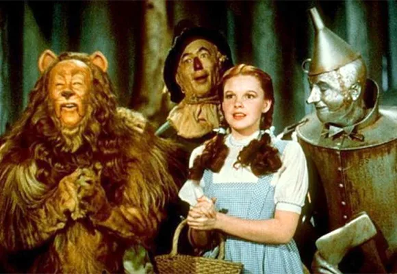 Four characters, including a lion, scarecrow, girl, and tin man, from "The Wizard of Oz" stand together in a forest.