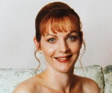 A smiling woman with reddish hair and pearl earrings sitting on a floral-patterned sofa.
