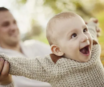 A smiling baby with open arms, held by an adult in a blurred outdoor setting.