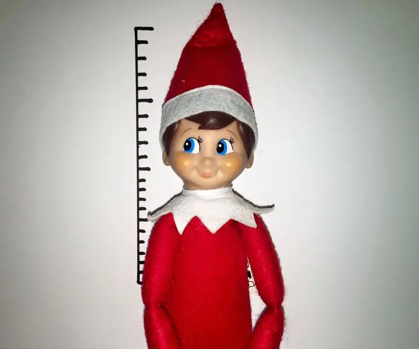 Elf on the Shelf doll in red outfit with hat, posed against a height chart.