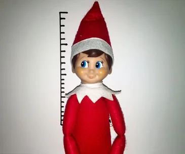 Elf on the Shelf doll in red outfit with hat, posed against a height chart.
