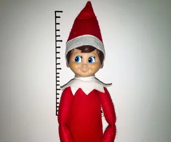 Elf on the Shelf doll in red outfit with hat, posed against a height chart.
