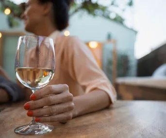 A person holding a glass of white wine with red nail polish, sitting outdoors at a table.