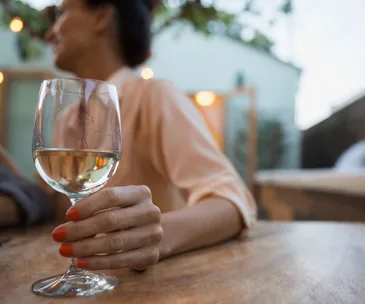 A person holding a glass of white wine with red nail polish, sitting outdoors at a table.