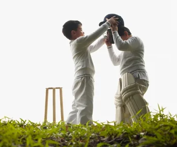 Two young boys in cricket gear, one helping the other with a helmet, near a wicket on grassy ground.