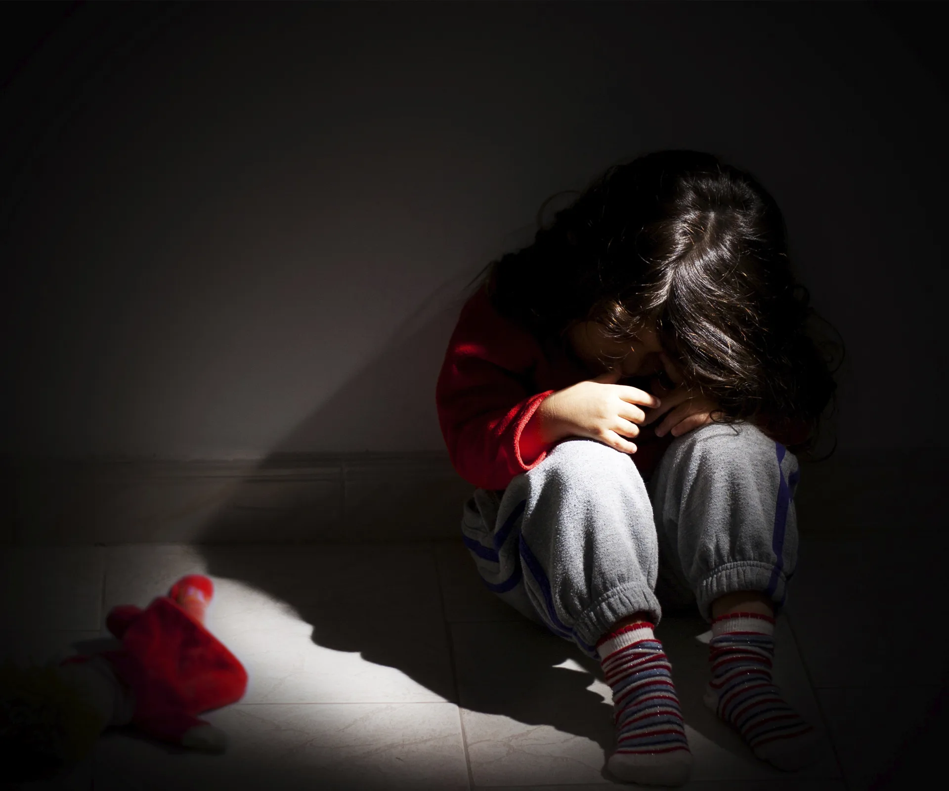 Child sitting alone in shadowed room, head in arms, with a toy doll on the floor beside them.