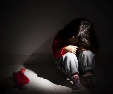Child sitting alone in shadowed room, head in arms, with a toy doll on the floor beside them.
