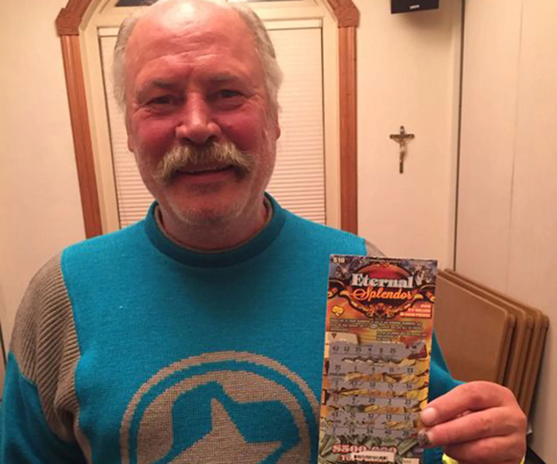 Man smiling while holding a winning scratch lottery ticket in a room with a crucifix on the wall.