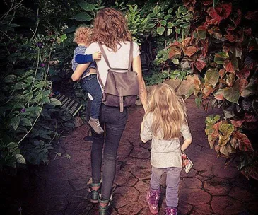 A woman with a backpack walks down a garden path holding a toddler, followed by a young girl carrying a book.