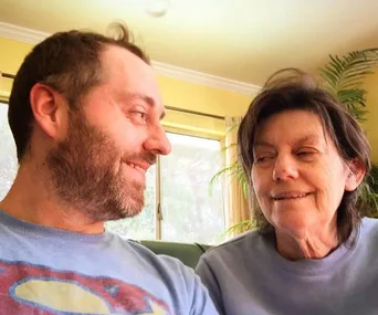 A smiling man sits with a woman inside a bright room, sharing a warm moment together.