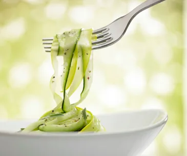 Fork twirling zucchini noodles in a white bowl against a soft green background.