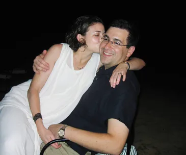 A woman kisses a smiling man on the cheek while sitting on his lap in a candid moment.