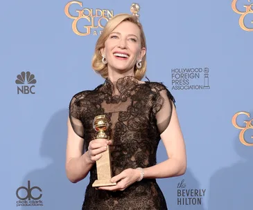 Actor in lace dress holds Golden Globe award, smiling in front of the press backdrop.