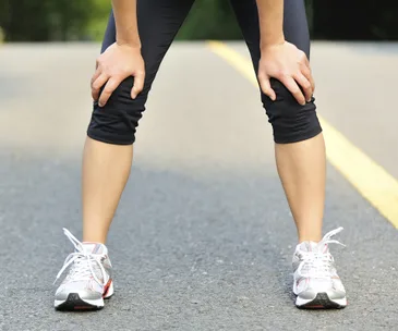Runner pausing on a road, legs bent, hands on knees, wearing black leggings and white sneakers.
