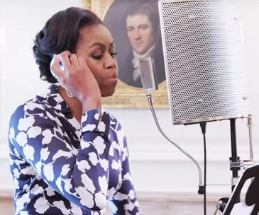 Michelle Obama, wearing a floral blouse, stands at a microphone in a recording studio setting.