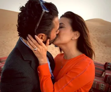 A couple shares a kiss in a desert setting, the woman wearing an orange dress and displaying an engagement ring.